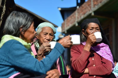 Three elderly women wearing sweaters are sipping tea; the sky behind them is a bright blue.